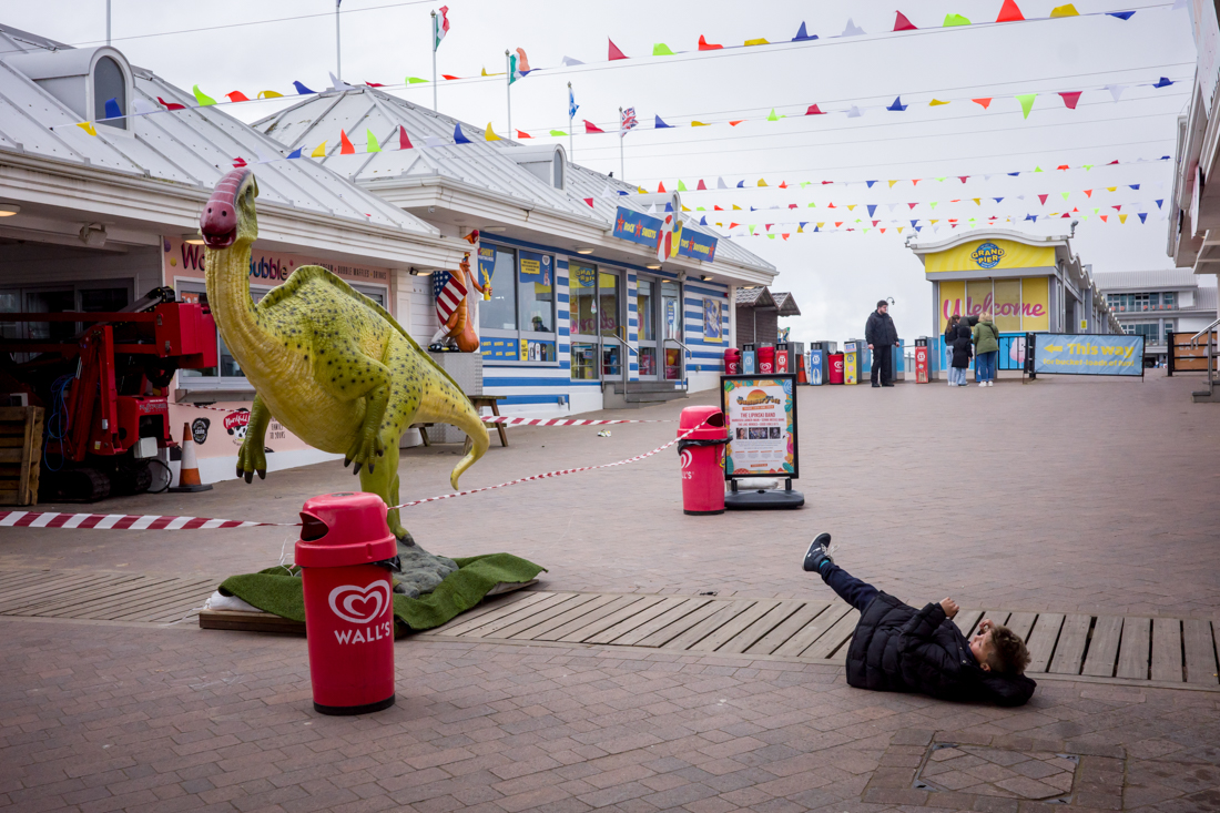Candid street photography of child falling over near a dinosaur on the Grand Pier in Weston-superMare, UK.