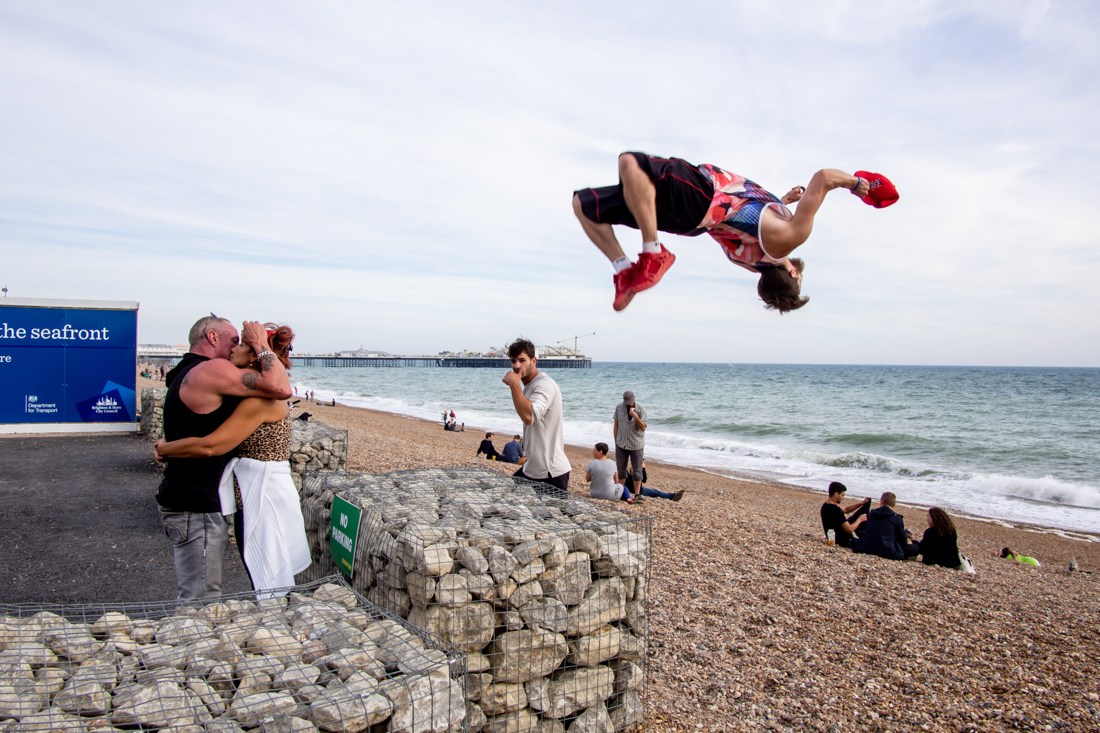 Street photography on Brighton seafront showing a man mid-air performing a backflip in front of the beach, while a couple kiss passionately and groups of people relax by the sea.
