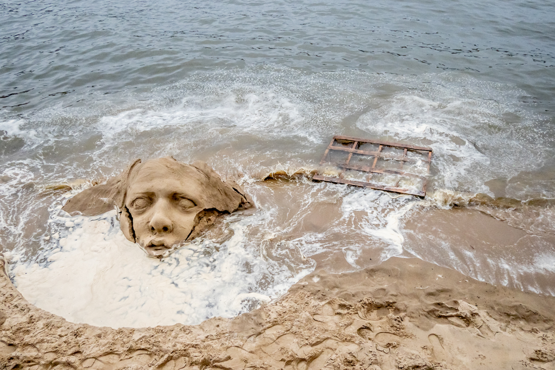 Candid street photography of a sand sculptured face seemingly drowning in the River Thames, in London.