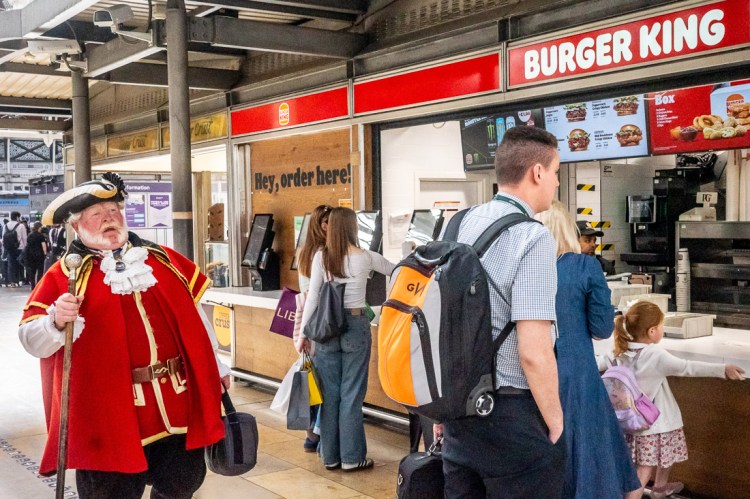 Candid street photography of a man in regal costume by the Burger King kiosk in London Paddington station.