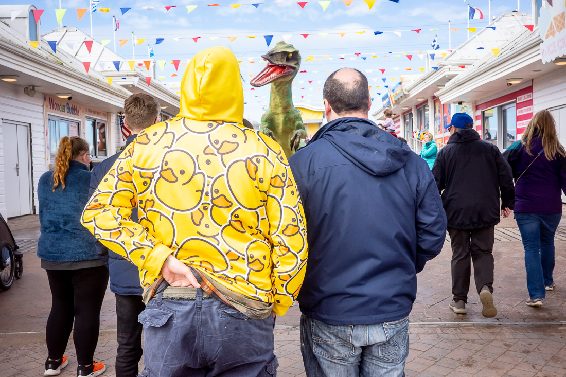 Candid street photography of a dinosaur roaring at a man in a duck hoodie, on the Grand Pier in Weston-super-Mare.