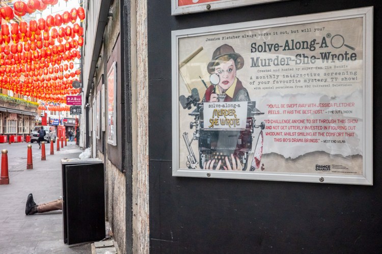 Candid street photography of legs sticking out from behind an electricity box, and a murder sigh, in Chinatown, London.