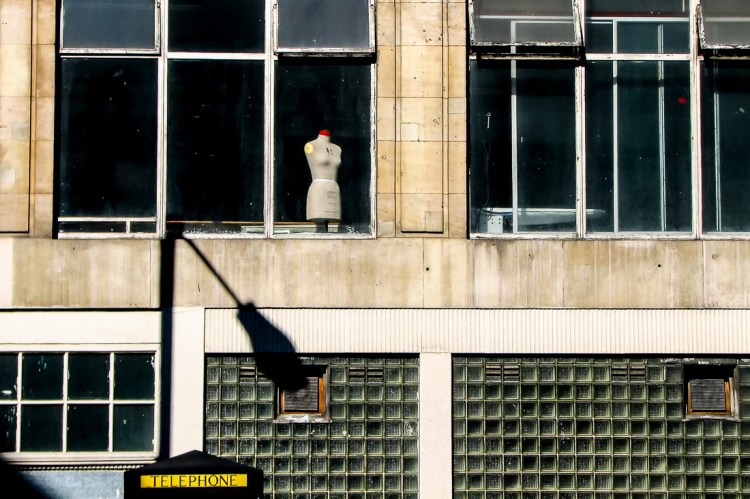 Candid street photography taken in the City of London on 13 July 2008, featuring a solitary mannequin in a window above a brutalist building facade, evoking themes of loneliness and urban isolation.