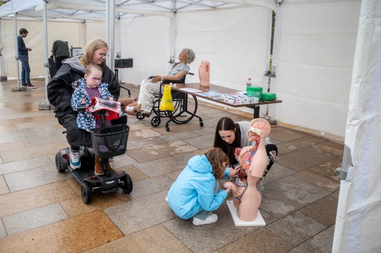 Street photography scene at the Italian Gardens in Weston-super-Mare showing a mother and daughter on a mobility scooter observing a public science education stall. Nearby, a woman and young girl interact with a human anatomy model, while medical training mannequins sit in the background under white event tents. The image captures curiosity, accessibility, and community engagement in a rainy urban setting.