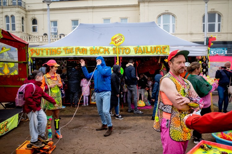 Vibrant street photography from the Whirligig Festival in Weston-super-Mare, capturing a colourful carnival stall with the sign “Putting the Fun Back into Failure.” Performers in eccentric, neon outfits interact with festival-goers, including a child on a balance game and a man using a quirky breathing tube contraption. The playful chaos and rain-soaked ground add to the surreal, joyful energy of the scene.