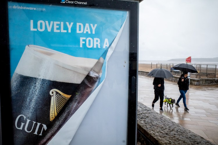 Candid street photo taken on the seafront in Weston-super-Mare on a rainy day, showing two people walking dogs under umbrellas. In the foreground, a humorous Guinness advert reads “Lovely day for a…” against the irony of the grey, wet weather.