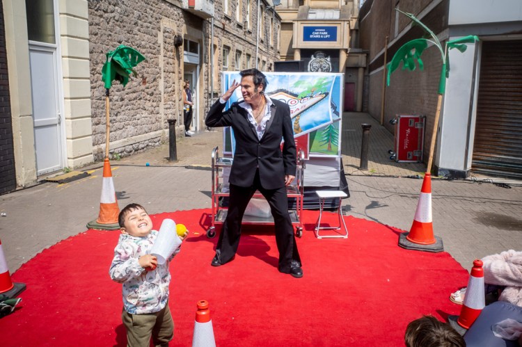 Candid street photography taken at the Whirligig Festival on Regent Street in Weston-super-Mare, featuring a lively performance by Elvis Bond on a red carpet stage, with a joyful child in the foreground holding traffic cones and laughing in the sunshine.
