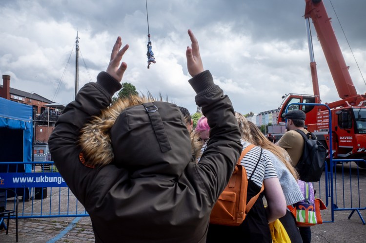 Candid street photography taken at the Harbourside in Bristol, showing a crowd watching a bungee jumper mid-air against a dramatic cloudy sky, with a young boy in a hooded coat raising their arms in the foreground.