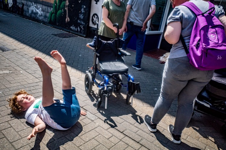 A candid street photography moment on the High Street in Weston-super-Mare, capturing a child playfully rolling on the pavement with legs in the air, beside an empty wheelchair and surrounded by passers-by. The scene is full of movement and everyday chaos, with urban textures, graffiti walls, and contrasting human interactions adding visual tension and humour.
