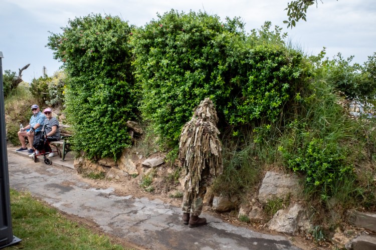 Candid street photography taken at Armed Forces Day on the Beach Lawns in Weston-super-Mare, showing a person in full camouflage gear blending into greenery while two amused spectators watch from a nearby bench.