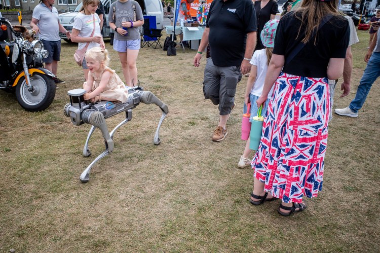 Candid street photography taken at Armed Forces Day on the Beach Lawns in Weston-super-Mare, featuring a young girl riding a robotic dog surrounded by families and Union Jack-themed outfits at a lively outdoor event.