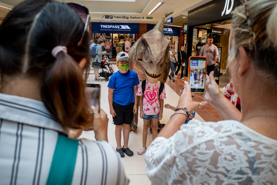 Street photo in Weston-super-Mare shopping centre with two children posing for photos, one in a dinosaur costume head and the other with face paint, while parents capture the moment on their phones.