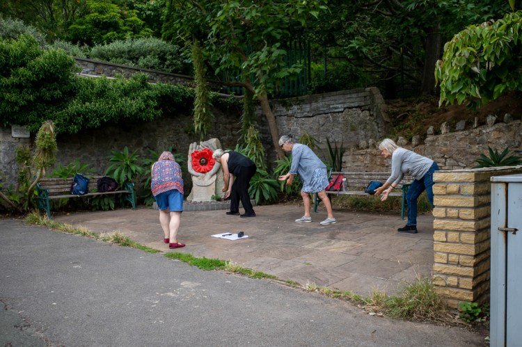 Street photo in Grove Park, Weston-super-Mare showing a small group of older women bending forward in unison near a hand sculpture with a red flower, during an outdoor activity.