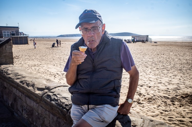 Street photo on Weston-super-Mare beach of an elderly man wearing a cap and glasses, eating an ice cream cone while leaning against a stone wall with sand and the sea in the background.