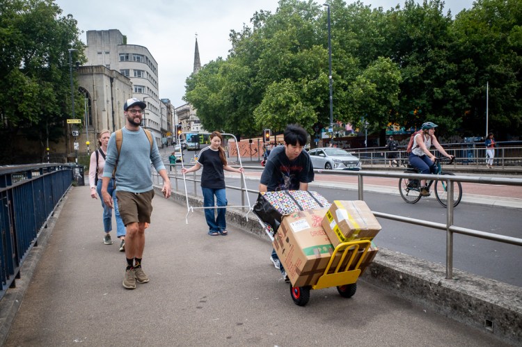 Street photography in Bristol capturing a busy urban moment with a man pushing a trolley stacked with cardboard boxes, another person carrying a metal frame, and pedestrians and cyclists passing by on the pavement and road.