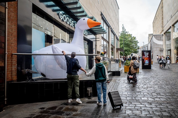 Street photography in Bristol showing two people holding hands while one touches a giant inflatable goose outside a shopfront, with shoppers and passersby in the background on a wet city street.