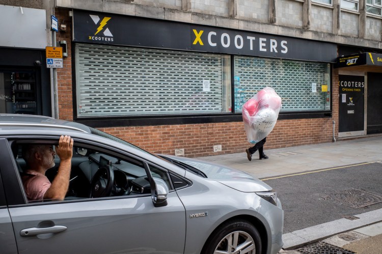 Street photography in Bristol showing a man walking past closed shop shutters while carrying a large bundle of colourful balloons, partly obscuring his body, as a driver looks on from a nearby car.