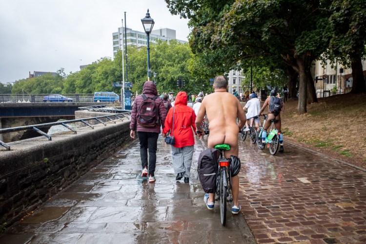 Street photography in Bristol capturing the World Naked Bike Ride, with cyclists riding nude along a riverside path on a wet day, passing pedestrians in coats and rain jackets.