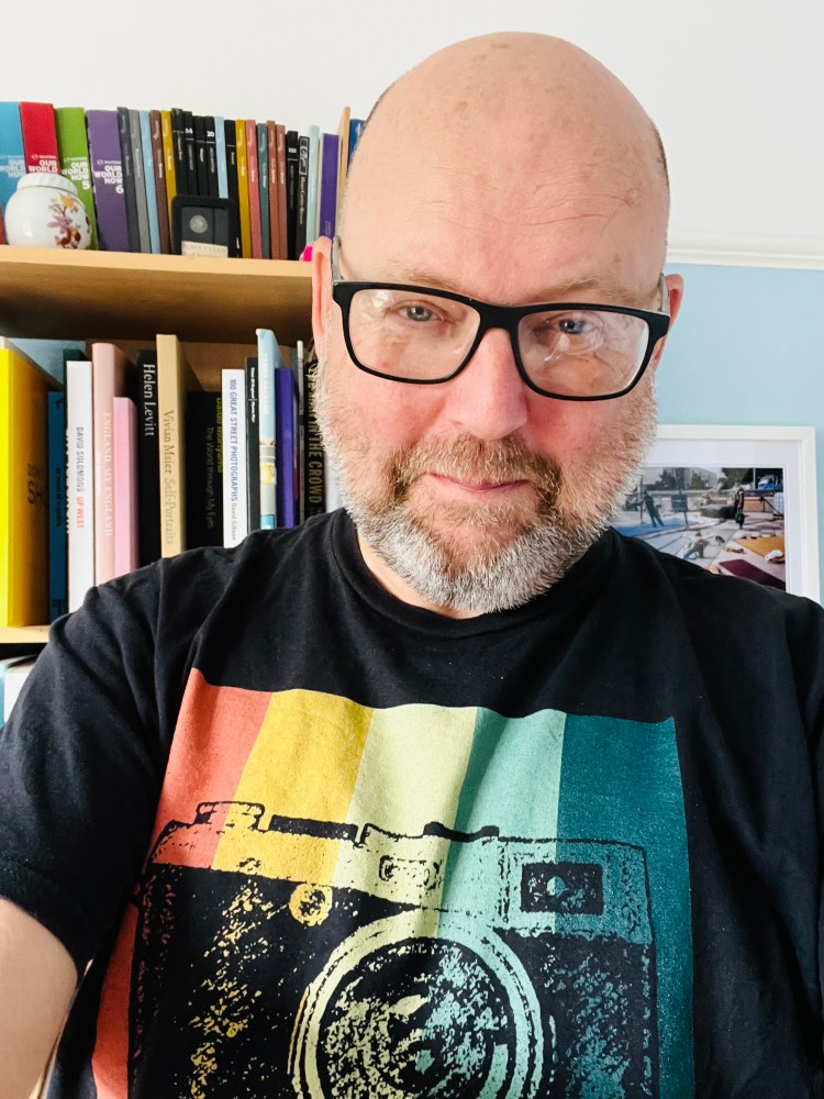Portrait of Darren Lehane wearing glasses and a black T-shirt with a colourful vintage camera design, standing in front of a bookshelf filled with photography books.
