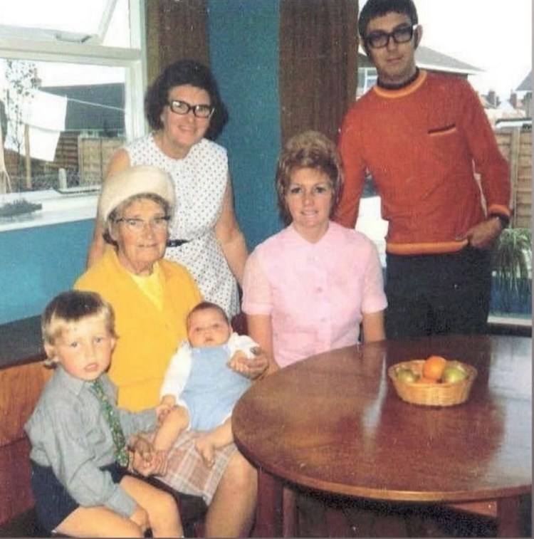 Family photo in the 1970s. At the front, I am sat next to my grandmother who is holding my younger brother. Behind us, my Nan stands in a white polka dot dress, my mother in a pink blouse sits at the table, and my father stands at the back in a red sweater.
