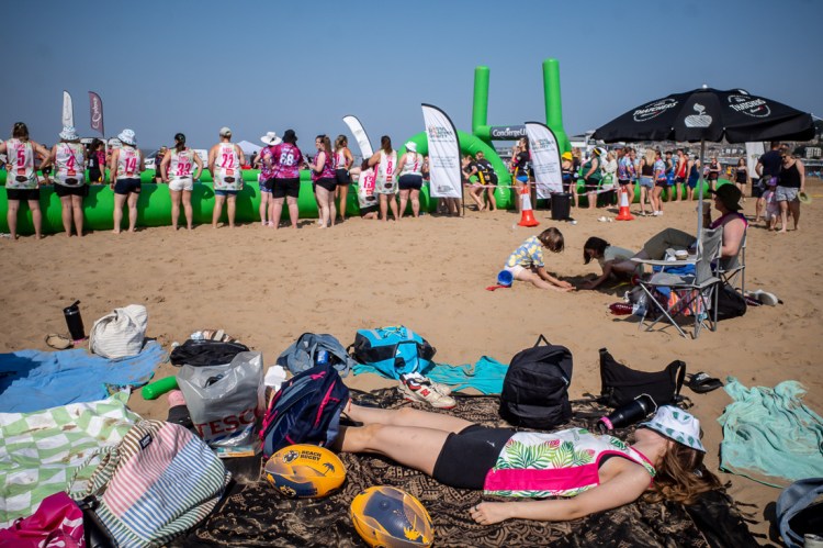 Street photography in Weston-super-Mare capturing beach rugby players and spectators, with a woman lying on a blanket resting in the foreground and teams preparing for matches on the sand behind her.