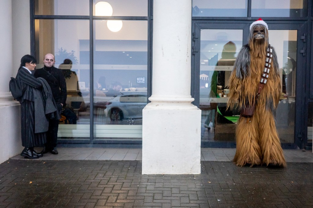 Candid street photograph outside the Wintergardens in Weston-super-Mare showing two people dressed in gothic-style outfits standing by a window while someone in a Chewbacca costume with a Santa hat waits nearby.