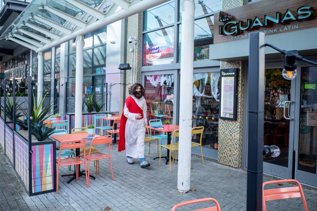 Candid street photography in Bristol showing a man dressed in a biblical-style costume walking past colourful outdoor seating at Las Iguanas on the Harbourside.