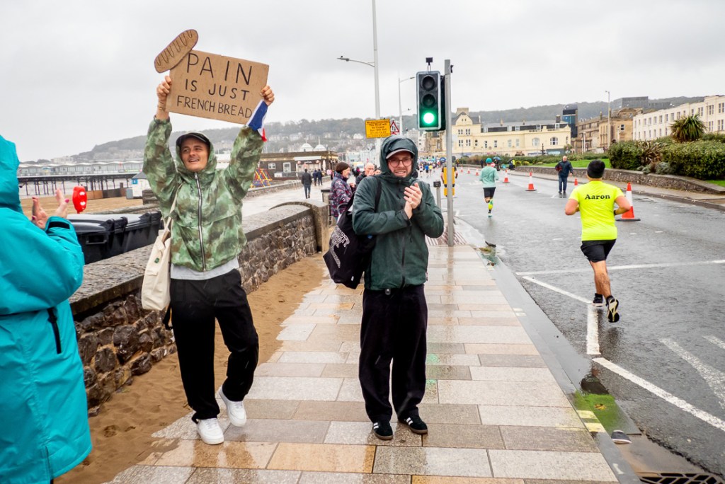 Candid street photo in Weston-super-Mare during a rainy marathon, showing a cheerful spectator holding a “Pain is just French bread” sign and another clapping, while runners pass along the seafront promenade on a wet, grey day.