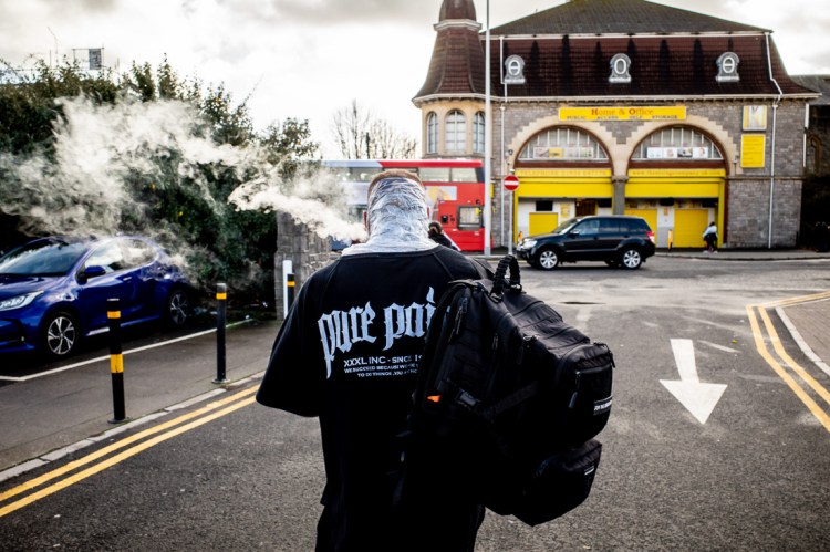 Candid street photography in Weston-super-Mare showing a person walking away while exhaling vapour, wearing a black ‘Pure Pain’ T-shirt and carrying a large backpack, with a red bus and local buildings in the background.