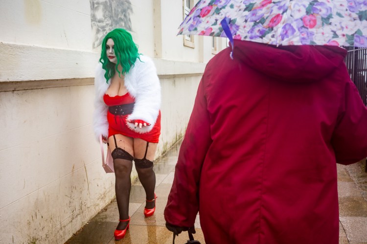 Candid street photograph outside the Wintergardens in Weston-super-Mare showing a person in a bright green wig, red dress and white faux-fur coat walking past someone holding a floral umbrella on a rainy day.