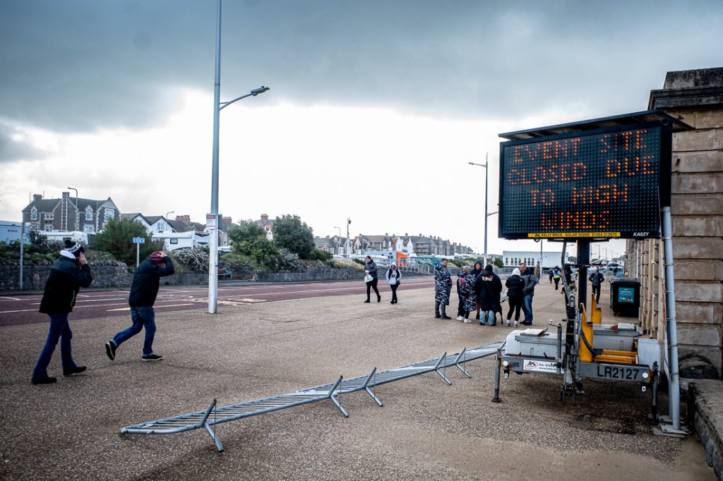 Candid street photo in Weston-super-Mare showing people battling strong winds on the seafront, with a digital sign reading “Event site closed due to high winds” and a fallen metal barrier on the promenade under dark storm clouds.