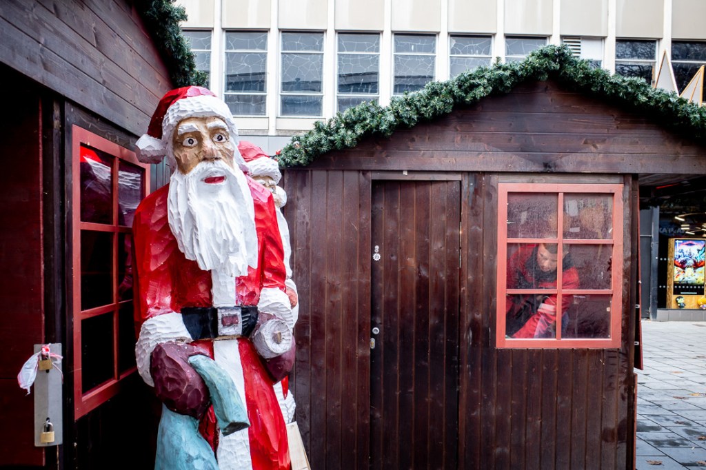 Candid street photograph at the Broadmead Christmas Market in Bristol showing a large carved Santa statue beside a wooden hut with a worker inside preparing for the festive stalls.