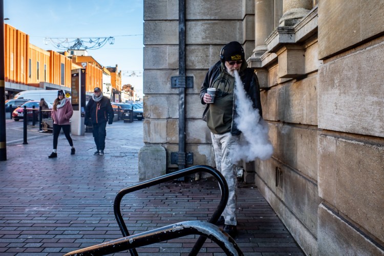 Candid street photograph on Abington Street in Northampton showing a man exhaling a large cloud of vapour while holding a coffee cup, with pedestrians walking in the background on a bright winter day.