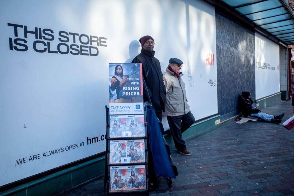 Candid street photograph on Abington Street in Northampton showing two Jehovah Witness standing beside a literature stand with ‘Coping With Rising Prices’ displayed, in front of a closed H&M store, while another person sits on the pavement in the background.