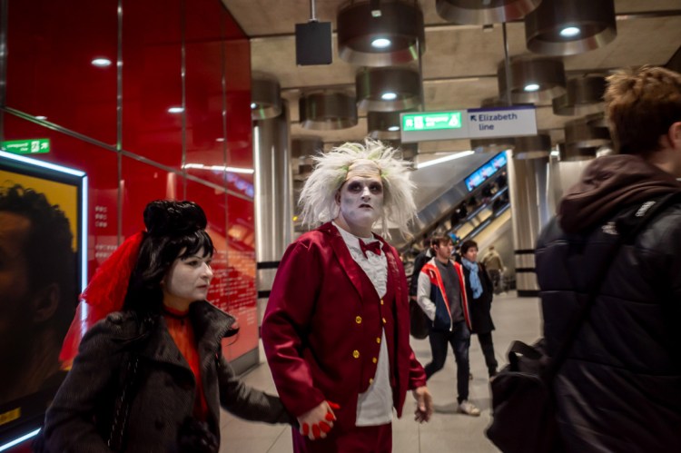 Candid street photograph inside Tottenham Court Road Underground station showing two people in dramatic Halloween-style costumes walking through the brightly lit concourse, with commuters in the background and Elizabeth line signage overhead.