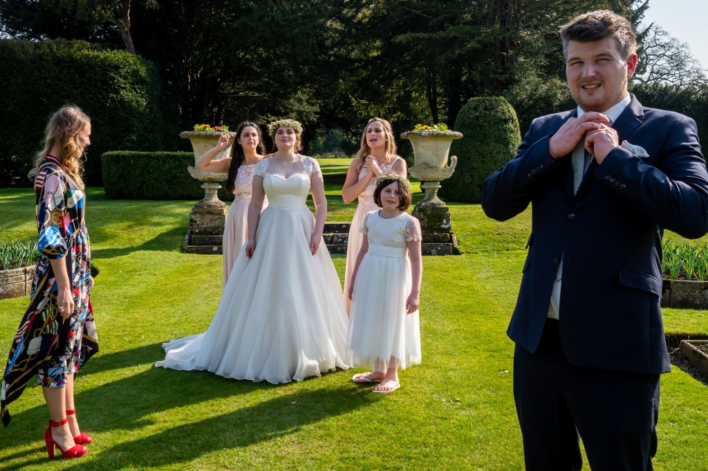 A wedding guest rearranges his tie, beside the bridal party, in the gardens at Grittleton House in Wiltshire.