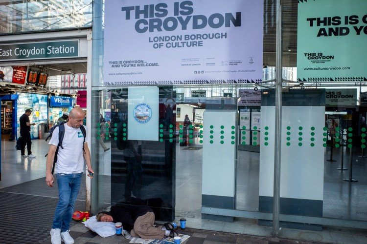 Candid street photography of a man sleeping rough under a sign for Croydon in London.
