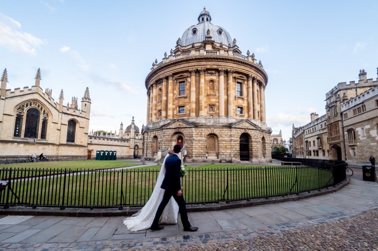 The bride and groom walk past the Radcliffe Camera during a wedding day in Oxford.