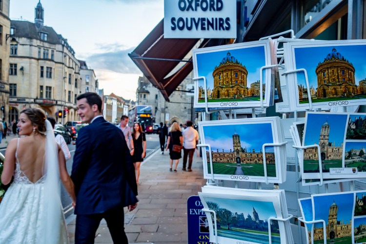 The bride and groom walk past some postcards during a wedding day in Oxford.