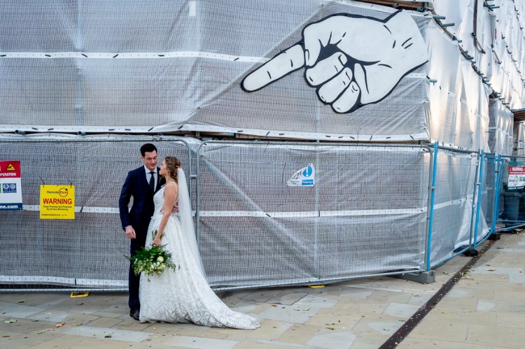 The bride and groom chat beneath a giant pointing hand and finger, on a wedding day in Oxford.