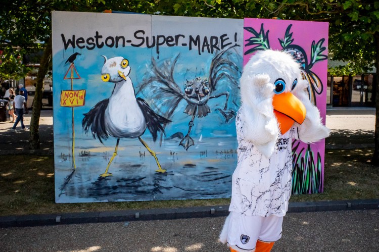 Candid street photography of a man dressed as a seagull in front of street art of a seagull in Weston-super-Mare, North Somerset, UK.