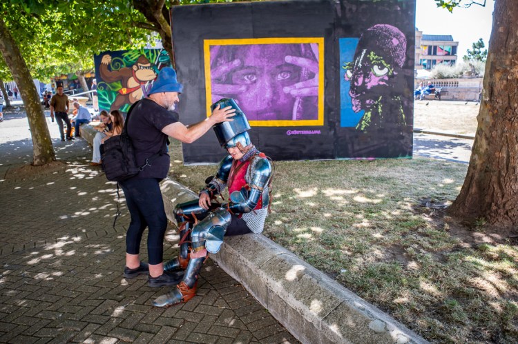 Candid street photography of a man in a suit of armour, having his helmet taken off in Weston-super-Mare, North Somerset, UK.