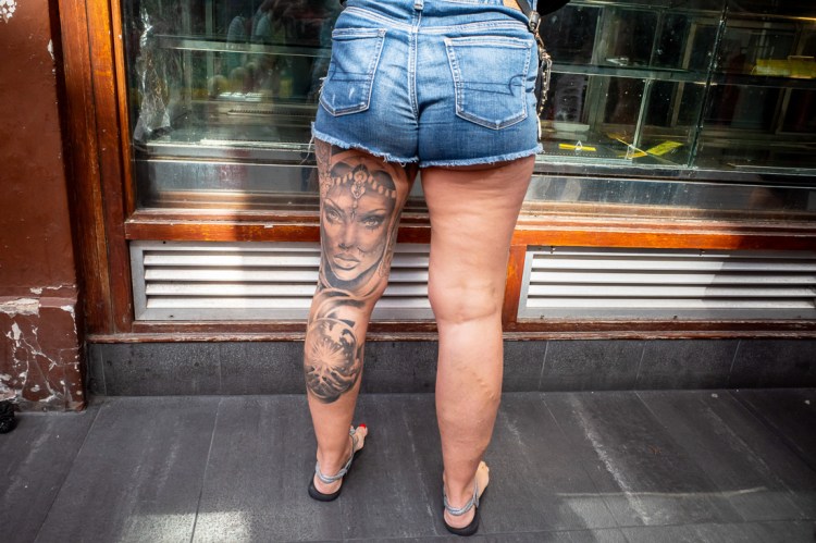 Candid street photography of a woman's face tattoo on the back of her leg in Chinatown, London.